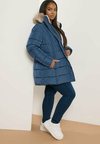 Blå pufferjacka med en pälsfodrad huva, med horisontella quiltade sektioner, öppen fram, och sidofickor, tillsammans med mörka jeans och vita sneakers.