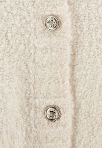 Texturiertes beige Kleidungsstück aus weichem, flauschigem Material mit zwei dekorativen silbernen Knöpfen mit kunstvollen Mustern.