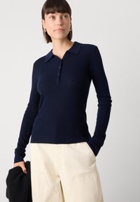 Polo shirt à manches longues côtelé bleu marine avec un col classique, une patte de boutonnage et une coupe ajustée, associé à un pantalon large beige.