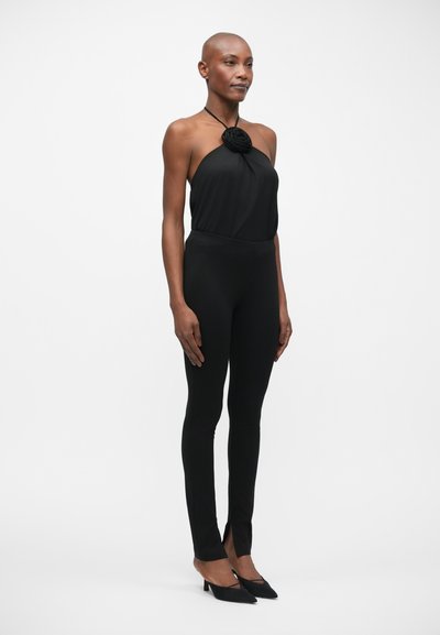 Zwarte halter jumpsuit met een rozen detail, aansluitend ontwerp, rekbare stof en taps toelopende benen met zijsplitten bij de enkels. Draagt met zwarte hakken.