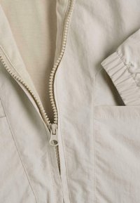 Veste beige avec fermeture éclair en plastique partiellement zippée, poignet élastique visible sur la manche droite, et poche sur le côté inférieur droit.