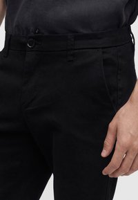 Close-up van een persoon die een zwarte broek met knoop en zijzakken draagt en een zwart ingestoken overhemd, hand rustend bij de dij.