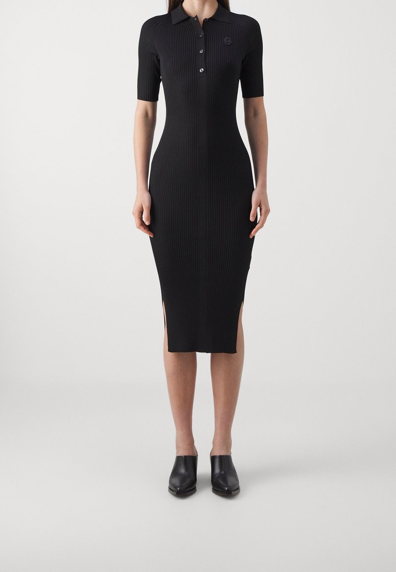 Femme portant une robe midi noire moulante en maille côtelée, avec des manches courtes, un col, des boutons sur le devant, des fentes latérales et des chaussures noires fermées.