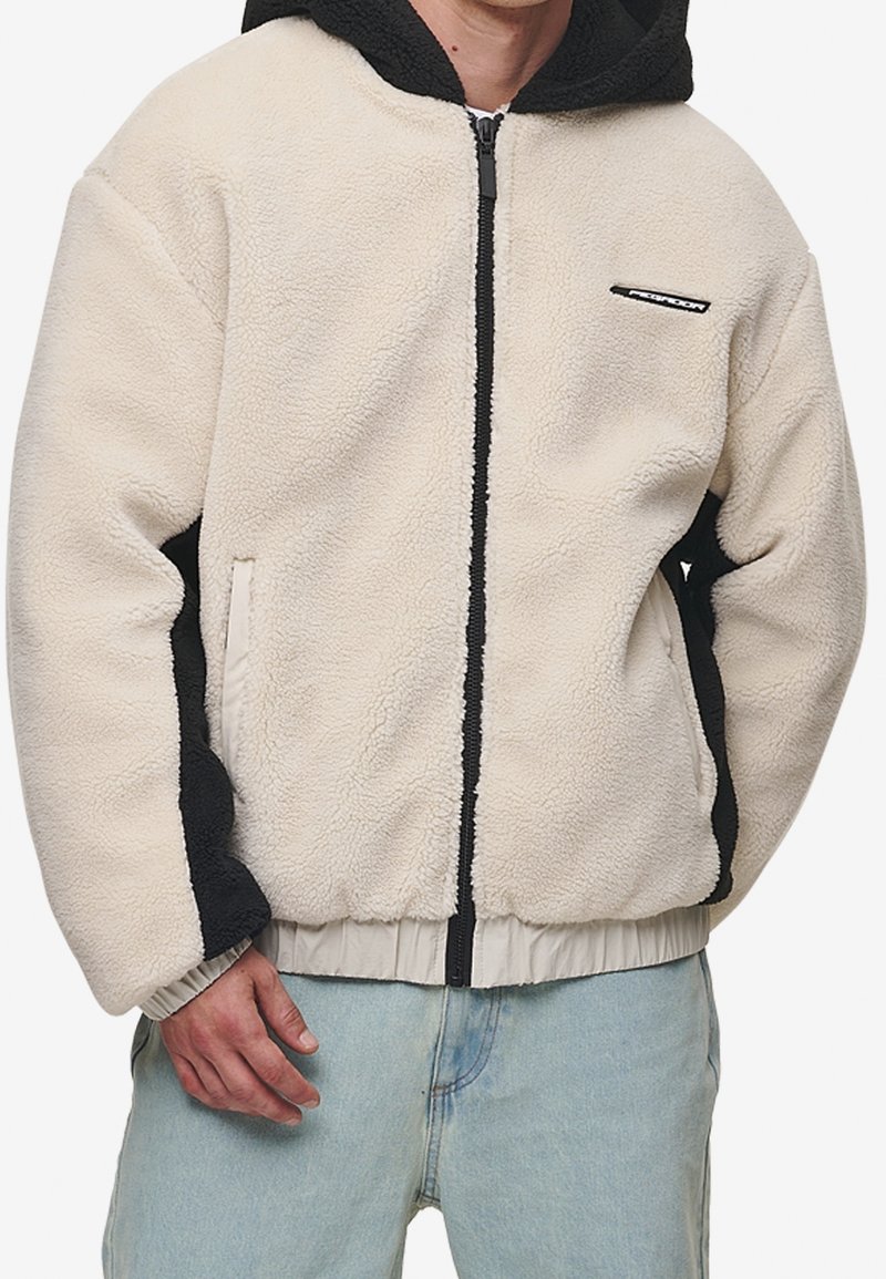 Giacca in pile crema con pannelli laterali neri, chiusura a zip, cappuccio e polsini elasticizzati. Presenta una texture morbida e un dettaglio di marchio minimale.