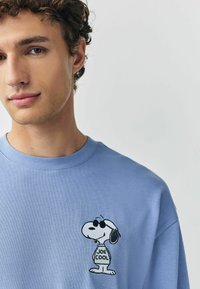 Svetlomodré bavlnené tričko s vyšívaným dizajnom Snoopyho. Postava má na sebe slnečné okuliare a na tričku je nápis "JOE COOL".