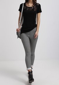 Kvinna i svart spetsblus, svart-vita rutiga byxor, svarta ankelstövlar och bär en grå axelväska.