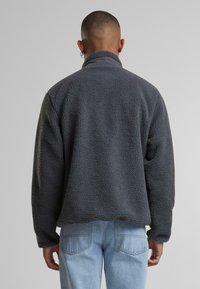 Grå fleecepullover med en tekstureret overflade, høj krave og afslappet pasform. Båret over lysblå jeans.