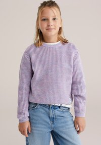 Joven con cabello rubio que lleva un suéter de punto lavanda, jeans azul claro y un cinturón blanco, de pie frente a un fondo neutro.