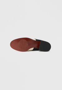 Semelle de chaussure noire à surface lisse, avec un dessous rouge, un détail de logo rond, et un talon noir texturé. Partie blanche à l'avant.