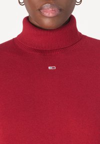 Maglione a collo alto rosso realizzato in tessuto a maglia con una texture a costine. Presenta un piccolo logo sul petto, che aggiunge un dettaglio di contrasto discreto.