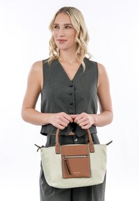 Borsa a mano color beige chiaro con dettagli in pelle marrone e tasca frontale con zip. Presenta manici doppi e una superficie liscia e impermeabile.
