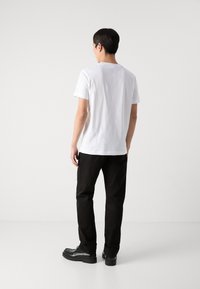 Uomo in piedi, di spalle, che indossa una semplice maglietta bianca, pantaloni neri e scarpe eleganti nere su uno sfondo bianco uniforme.