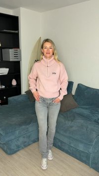 Femme debout dans une pièce portant un pull rose GAP, un jean gris et des baskets blanches, avec un canapé bleu et une planche de surf derrière elle.