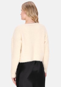 Kobieta z falującymi blond włosami nosi sweter w kolorze kremowym i czarną spódnicę, pokazana od tyłu na białym tle.