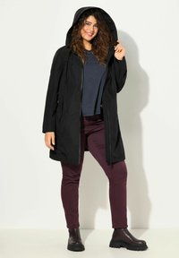 Chaqueta negra con capucha de textura suave, con cremallera en el frente y bolsillos laterales. Combinada con pantalones burdeos y botas negras gruesas.