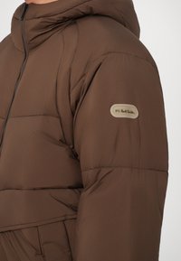 Barna puffdzseki kapucnival, cipzáras elülső résszel, steppelt textúrával és egy kis logóval a bal karon.