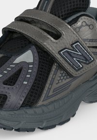 Παιδικό μαύρο και γκρι αθλητικό παπούτσι New Balance με λουράκια βέλκρο, επάνω μέρος από δίχτυ και σχέδια από συνθετικά υλικά, κοντινή όψη.