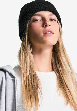 Beanie negru cu model în dungi purtat cu un hanorac gri cu fermoar. Persoana are părul lung, de un blond deschis, și o bluză albă minimalistă.