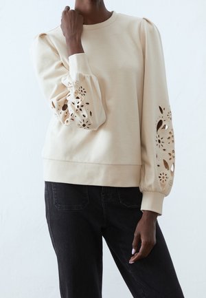 Persoon in een beige langemouwen top met bloemen uitsnijdingen op de pofferige mouwen en zwarte broek tegen een witte achtergrond.