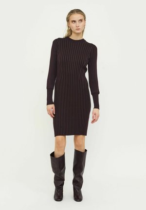 Mujer de pie con un vestido de punto negro acanalado hasta la rodilla y botas altas de cuero marrón oscuro sobre un fondo liso.
