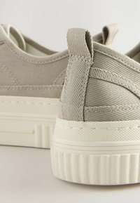 Sneakers beige in tela con superficie testurizzata, tallone rinforzato e suola in gomma bianca con motivo a righe. Nessun marchio visibile.