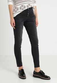 Černé džíny skinny s vysokým pasem, zapínáním na knoflík a cvočky na lemu. Nošeny s lesklými černými šněrovacími botami.