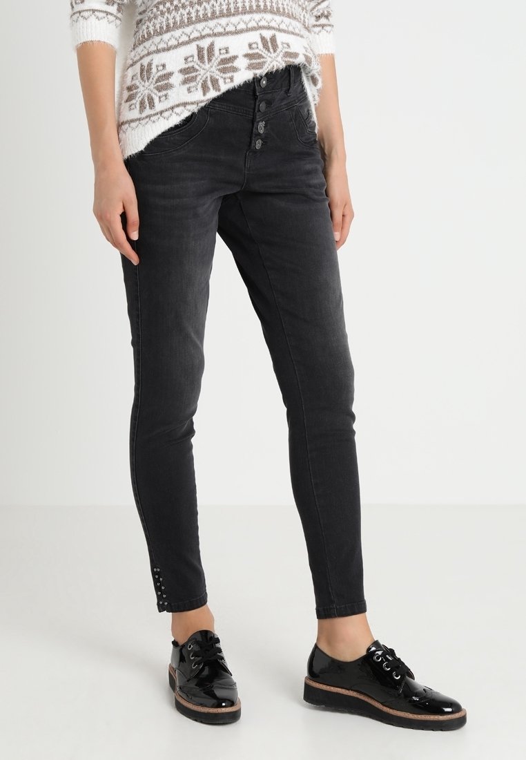 Černé džíny skinny s vysokým pasem, zapínáním na knoflík a cvočky na lemu. Nošeny s lesklými černými šněrovacími botami.