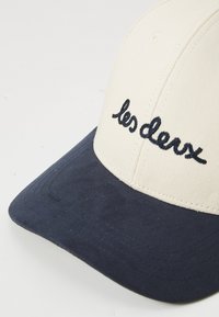Cream cap with a dark navy brim, featuring embroidered text "les deux" in black. Made of fabric with a smooth texture and curved visor.