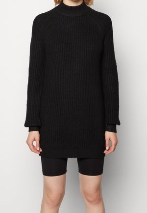 Persoon die een zwarte, lange mouwen, geribbelde gebreide sweaterjurk draagt met zwarte aansluitende shorts tegen een effen achtergrond.