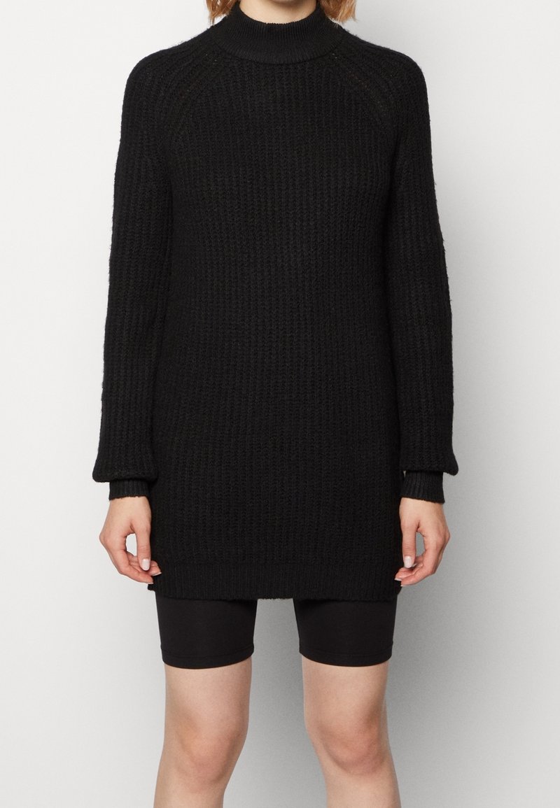 Personne portant une robe-pull noire en tricot côtelé à manches longues avec un short noir ajusté sur un fond uni.