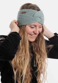 Fascia per testa lavorata a maglia di un blu pallido con un design attorcigliato e una piccola etichetta marrone. Tessuto testurizzato, indossata con capelli lunghi che scendono.