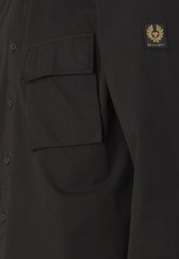 Black button-up shirt with a patch pocket and woven branding label on the left chest. Smooth textile with a structured fit.