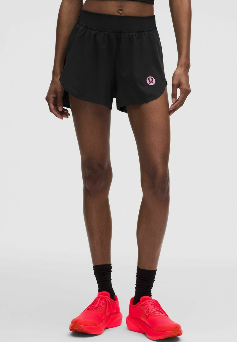 Athlet trägt schwarze Sportshorts mit pinkem Logo, schwarze Wadenhohe Socken und leuchtend rote Laufschuhe und steht vor grauem Hintergrund.