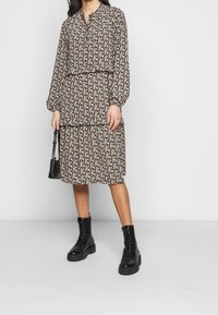 Černé květinové šaty s dlouhým rukávem, s nařaseným pasem a vrstveným designem; v kombinaci s černými šněrovacími botami a malou černou kabelkou.