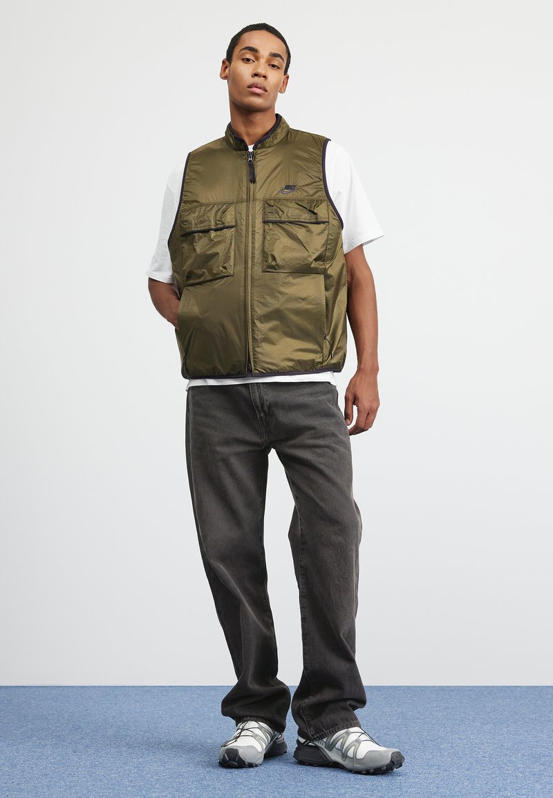 Jeune homme portant un gilet Nike vert olive sur un t-shirt blanc, un jean foncé et des baskets blanches-grises, se tenant contre un fond uni.