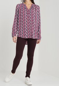 Blouse à manches longues avec un motif géométrique multicolore, dotée d'un col en V et d'une coupe ample, associée à des leggings sombres et des baskets blanches.