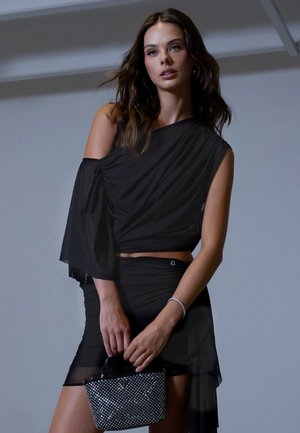 Femme aux longs cheveux bruns porte une robe asymétrique noire, tenant un petit sac à main noir clouté sur un fond gris uni.