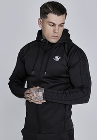 Czarna bluza z kapturem na zamek, wykonana z gładkiego materiału, z białym logo na klatce piersiowej oraz fakturowanym logo wzdłuż rękawów.