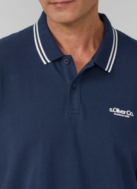 Marineblauwe polo met een gestructureerde stof, een knoopsluiting met twee knopen en een kraag met witte strepen. Merklogo aan de linkerkant.