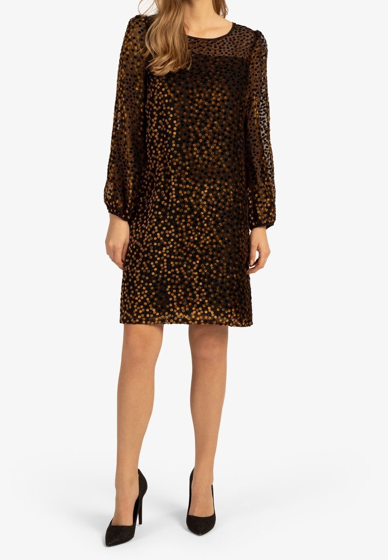 Femme portant une robe noire transparente à manches longues avec des pois dorés brillants et des talons hauts noirs, debout devant un fond blanc.