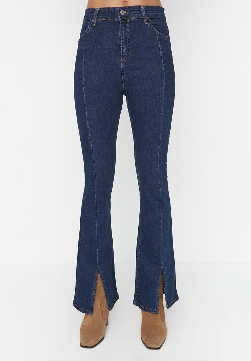 Jeans évasés bleu foncé avec ourlets fendus, taille haute et détails de coutures visibles. Associés à des bottines en daim marron clair.