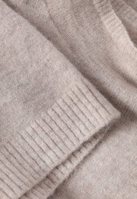 Világos bézs kötött pulóver közeli kép, puha tapintással, bordázott mandzsettákkal és sima testtel, finom színvariációkkal.