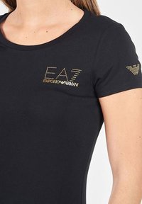 Juodas trumpomis rankovėmis marškinėliai su apvalia apykakle. Kairėje krūtinės pusėje yra auksinis EA7 logotipas, o dešinėje rankovėje - logotipo akcentas. Minkštas audinio paviršius.