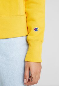 Felpa gialla con polsini a costine, texture morbida e un piccolo logo Champion sulla manica. Abbinata a jeans blu chiaro.