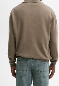 Brauner Strickpullover mit Polo-Kragen, ausgestattet mit langen Ärmeln. Getragen mit hellblauen Jeans mit einem lockeren Schnitt und Gesäßtaschen.