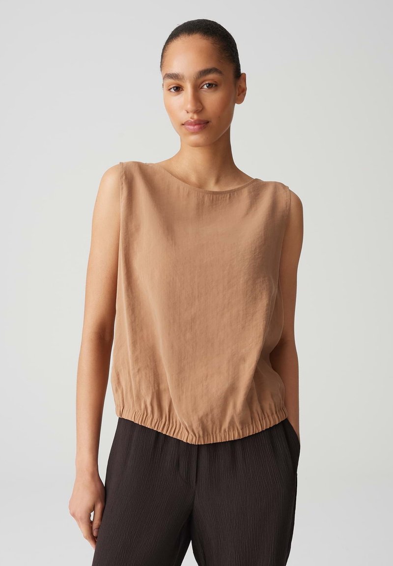 Top senza maniche color beige realizzato in tessuto leggero; presenta un colletto rotondo e un orlo elastico arricciato. Abbinato a pantaloni neri con texture.