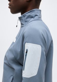 Πλάγια όψη ατόμου που φοράει ανοιχτό μπλε μπουφάν με ψηλό γιακά και τσεπάκι με φερμουάρ στο πάνω μέρος του μπράτσου, σε απλό φόντο.