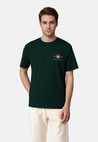 Dark green t-shirt made of cotton, featuring a small logo and text on the left chest area. Short sleeves and a crew neck design.