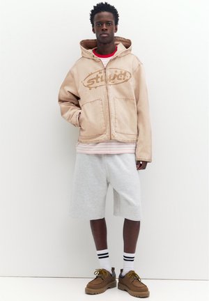 Młody mężczyzna ubrany w beżową bluzę z kapturem na zamek, koszulę w paski, jasnoszorty, białe skarpetki z czarnymi paskami oraz brązowe buty sznurowane, stoi przy białej ścianie.