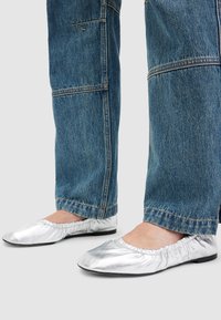 Picioare purtând balerini metalici argintii, asociați cu blugi de denim albastri cu cusături vizibile la tiv.